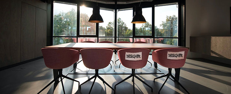 Foto di una sala riunioni con tavolo e sedie. Sullo schienale di due sedie è stato applicato un foglio con scritto "designer", per riservarle.